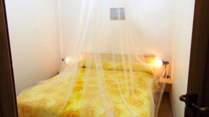 Une chambre avec un lit recouvert d’une couvre-lit à motifs floraux jaunes, une table de chevet avec une lampe et une petite fenêtre donnant sur un paysage agréable.