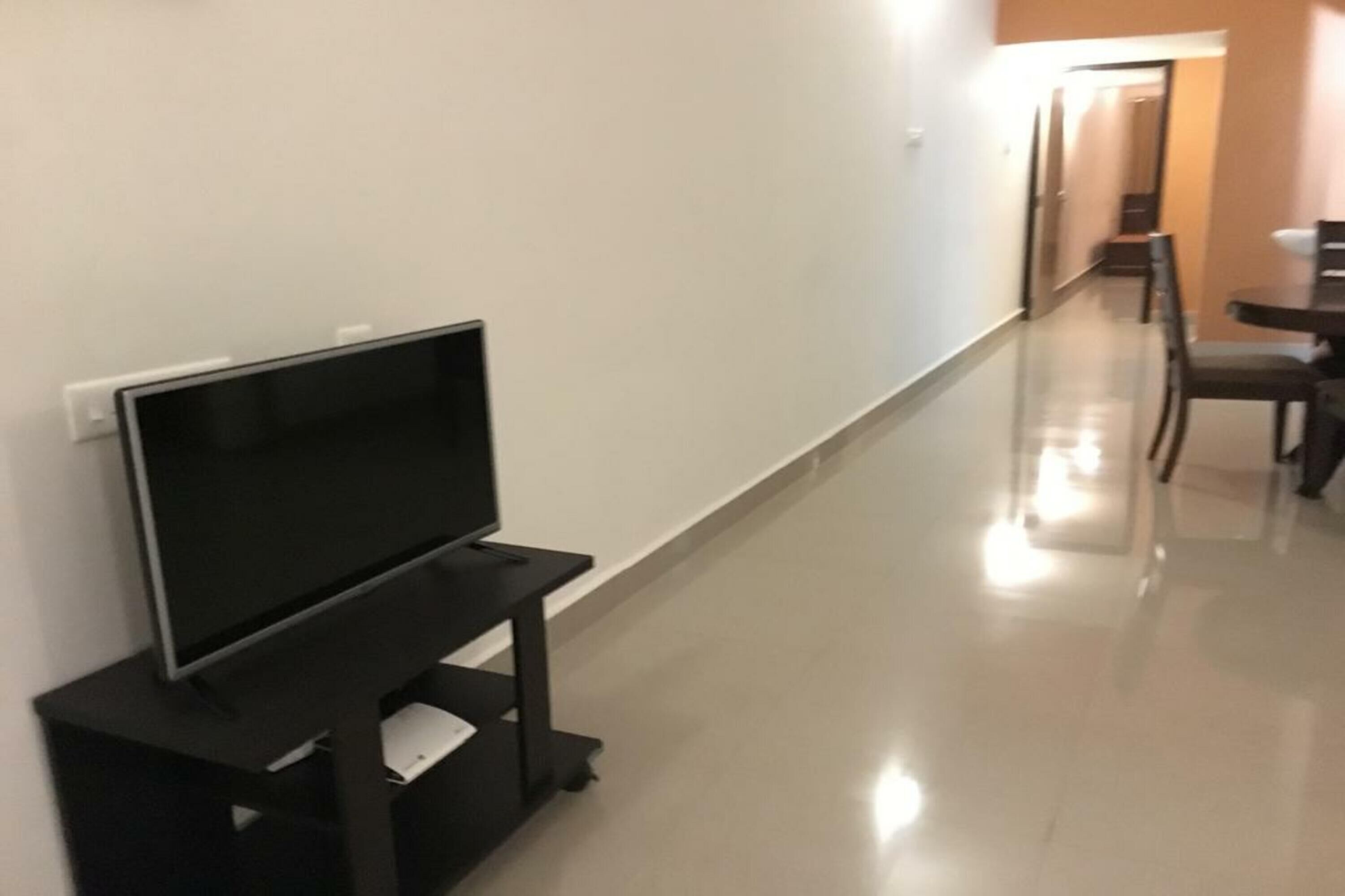 A hallway with a TV on a stand, a dining area with chairs and a table, and a door at the end.
