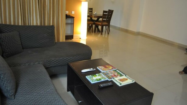 A living room with a sofa, a coffee table with magazines, a remote control, and a dining area in the background. - Jyothi Suites (Tiruchirappalli)