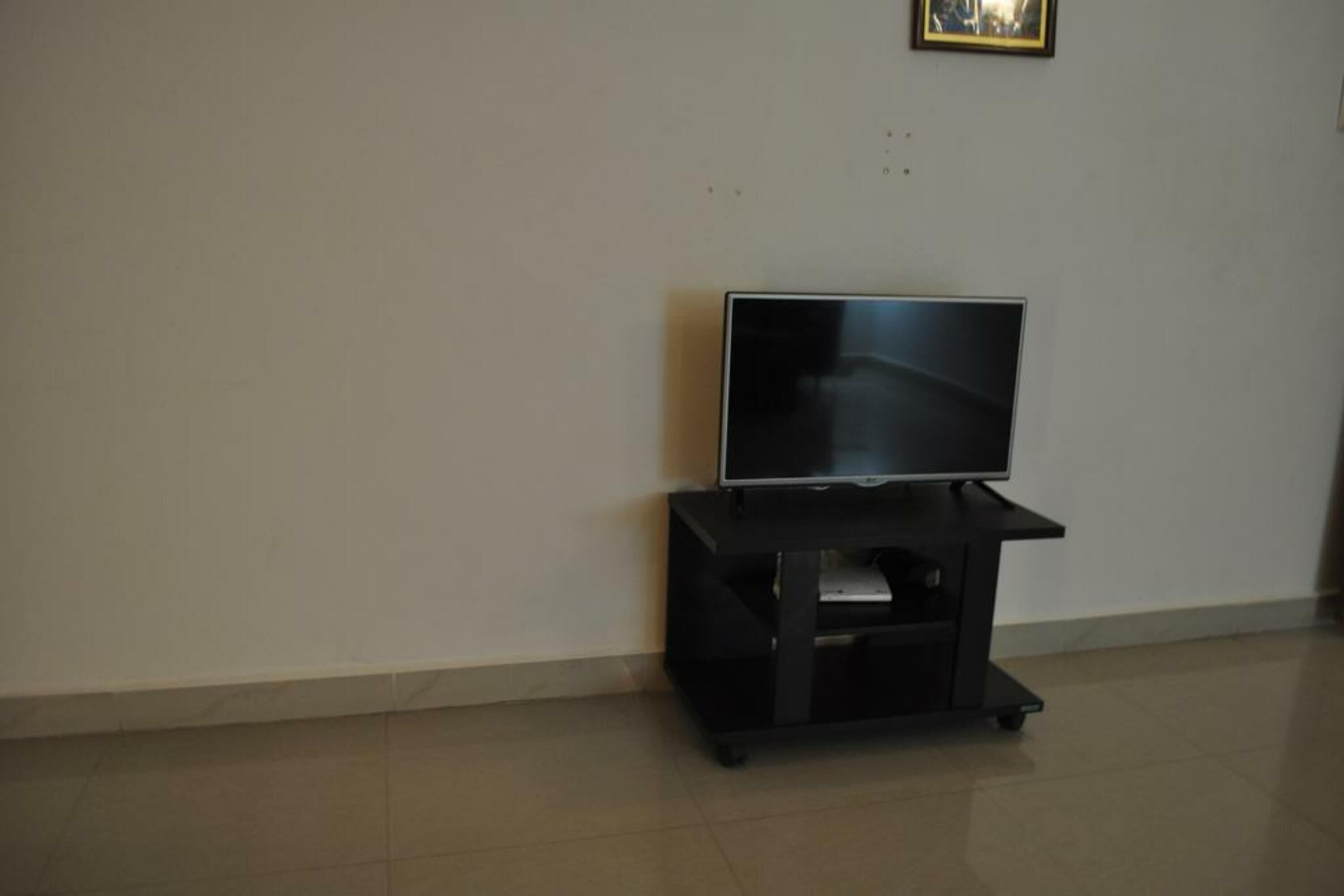A flat-screen TV on a black stand in a room with plain white walls and a tiled floor.