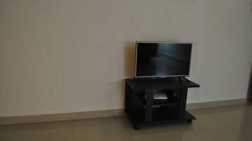 A flat-screen TV on a black stand in a room with plain white walls and a tiled floor.