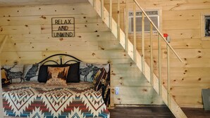 A cozy wooden cabin bedroom with a bed, a sign saying "RELAX AND UNWIND," and a staircase. -  Hocking Hills,Cozy Secluded Cabin,Minutes to Caves/WiFi/Pet Friendly! (McArthur)