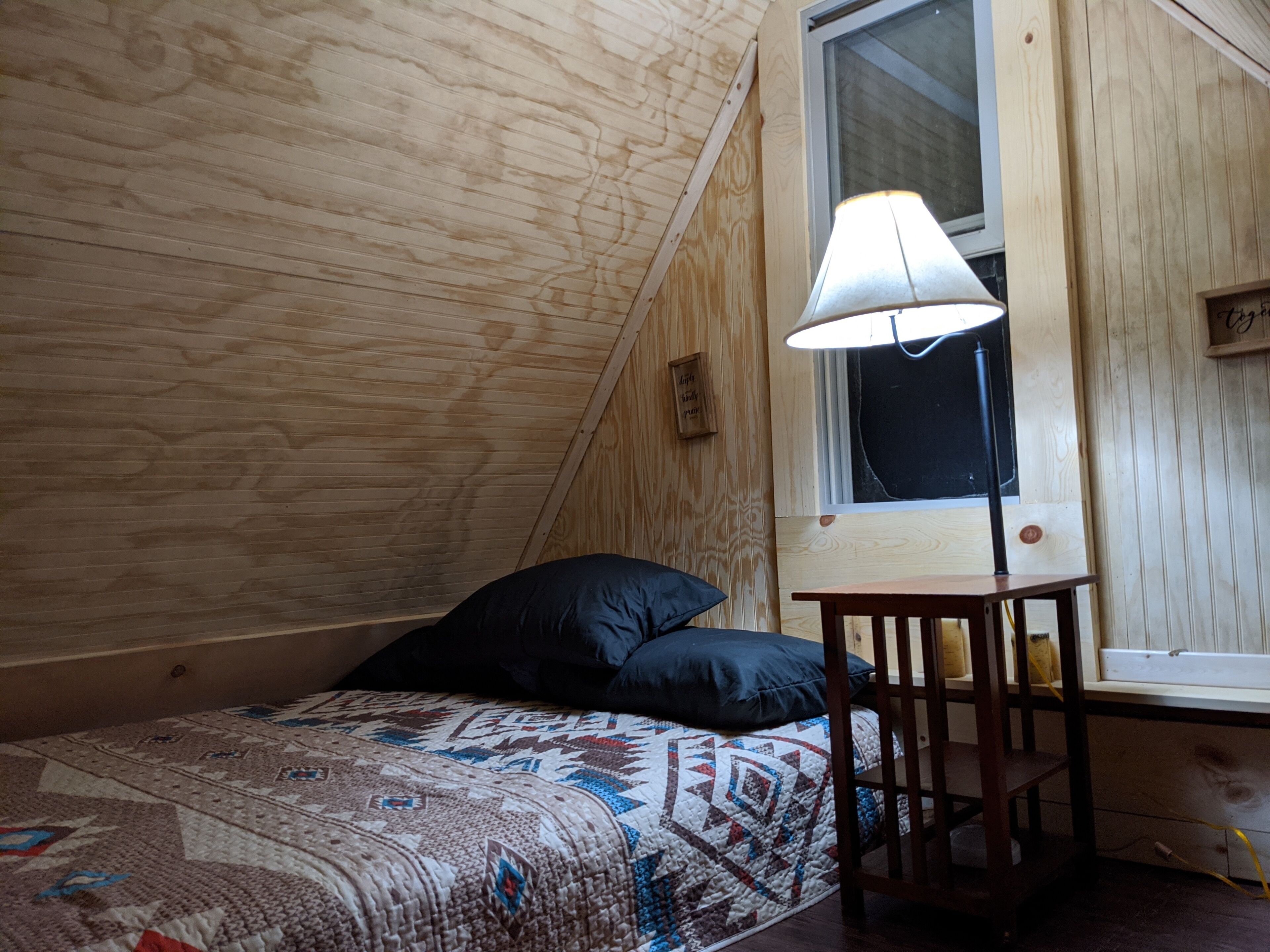 A cozy room with a sloped ceiling, a bed with a patterned quilt, a small table with a lamp, and a window with a view.