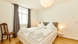 Una cama bien tendida con toallas dobladas, una silla de madera, una mesita de noche con lámpara y una ventana con cortinas.