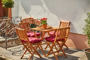 A wooden outdoor table and chairs with pink cushions, a potted plant with red flowers, and a metal railing.