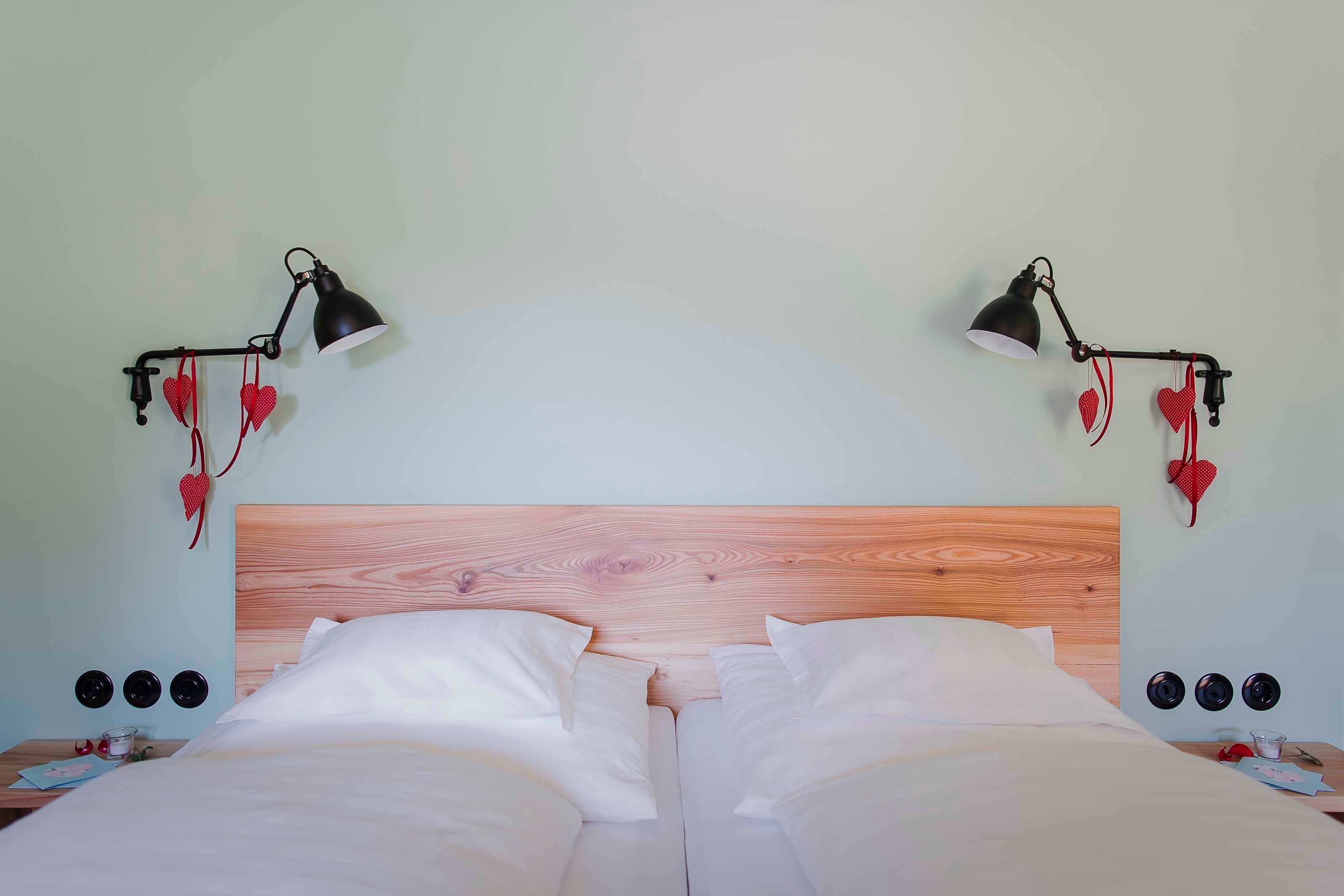 Un lit double avec du linge de lit blanc, deux tables de chevet avec des lampes et des décorations en forme de cœur accrochées au mur.