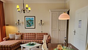 A living room with a checkered sofa, a white coffee table, a wooden dining table with a glass jar and bottles, a chandelier, and a framed painting on the wall.