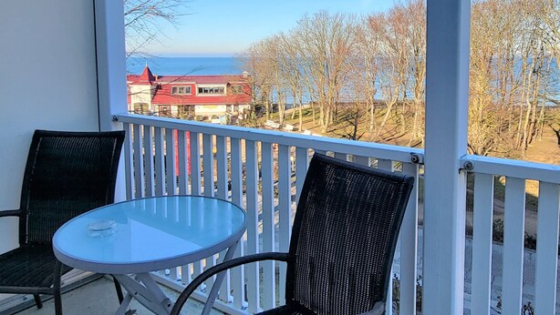 A balcony with a glass table and two wicker chairs, overlooking a residential area with houses and trees.