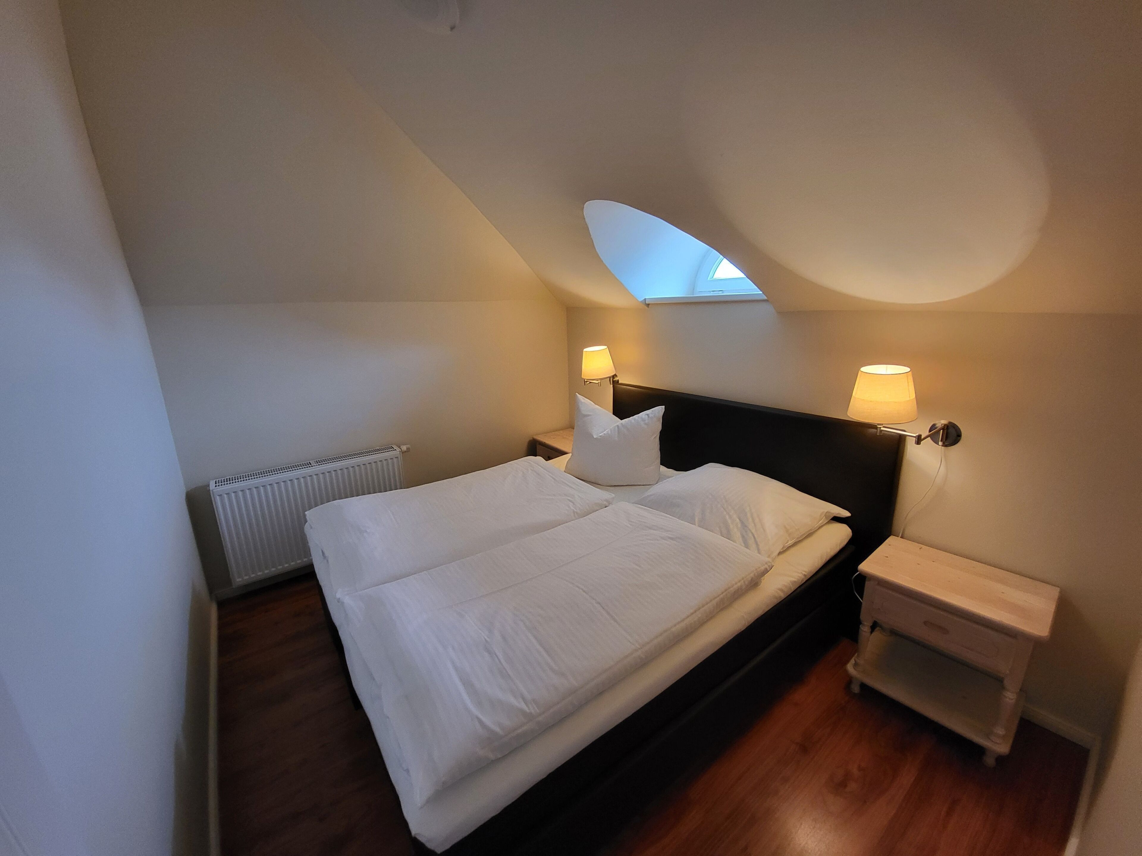 A bedroom with a bed, two bedside lamps, a nightstand, a radiator, and a skylight.