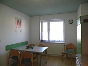 Une pièce comprenant une table à manger et des chaises, une fenêtre avec des stores, un radiateur et une horloge au mur.