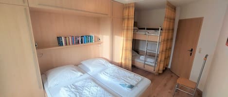 A bedroom with a large bed, a bunk bed, wooden shelving with books, and a small chair.