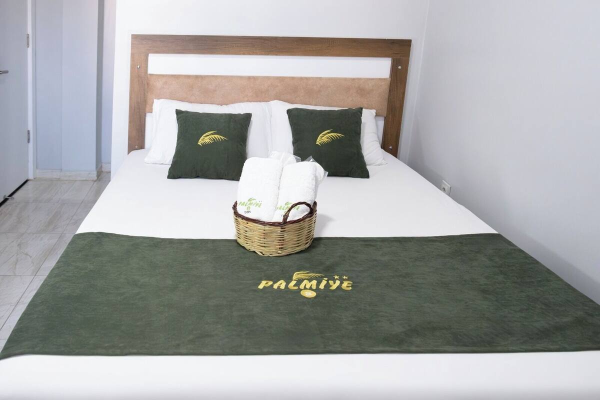 A neatly made bed with a green blanket, two pillows with a leaf design, and a basket of towels.