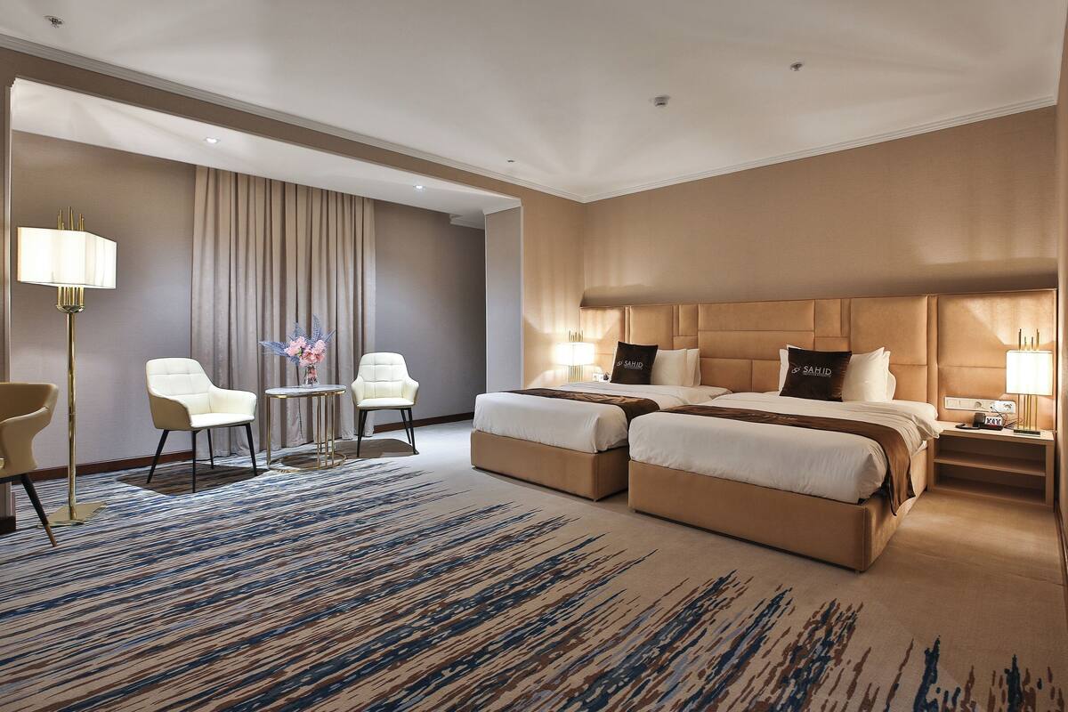 A hotel room with two beds, a sitting area with a table and chairs, and a large window with curtains.