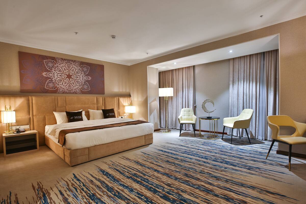 A hotel room with a large bed, two bedside tables, a desk with two chairs, a large abstract painting, and a view of the city through the window.