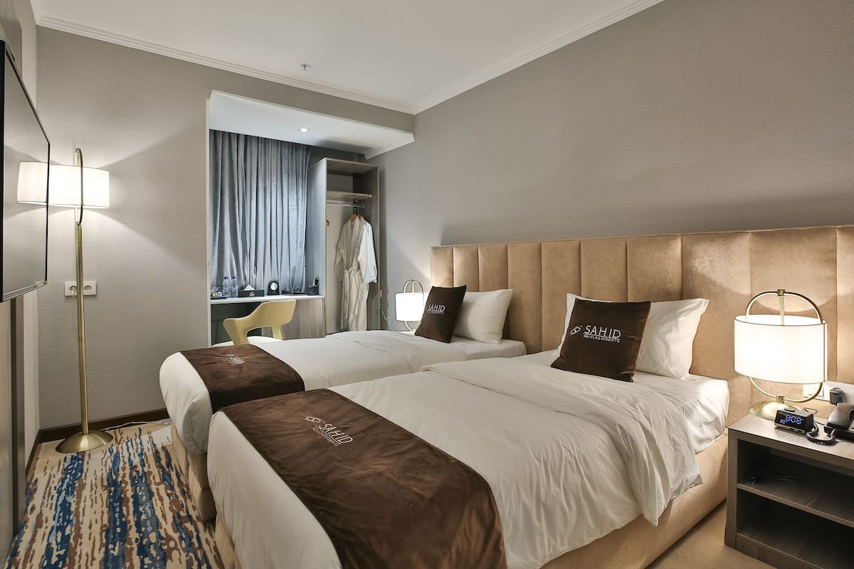 A hotel room with two beds, a bedside table with a lamp, a wardrobe, and a mirror.
