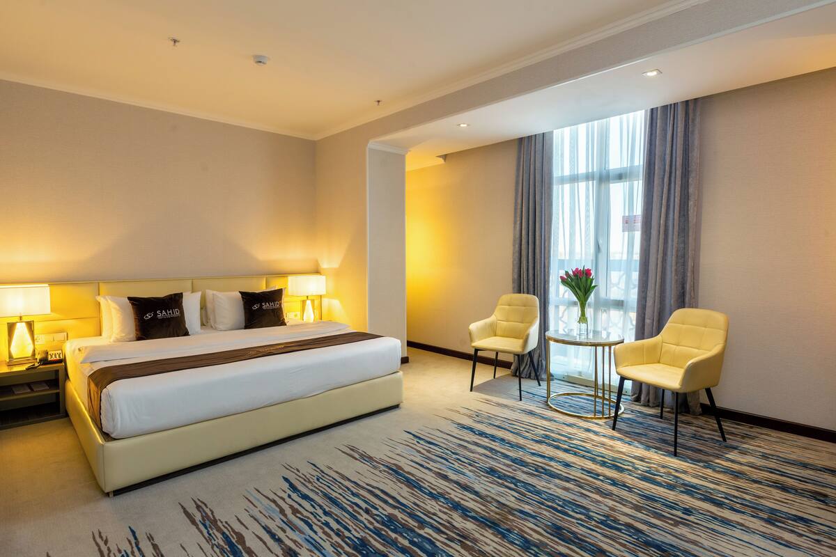 A hotel room with a large bed, two chairs, a small table, and a window with curtains.