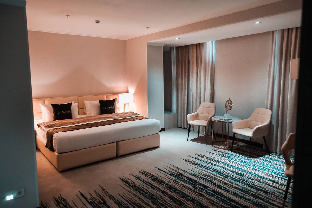 A modern hotel room with a large bed, a small table with chairs, and a view of the city through the window.