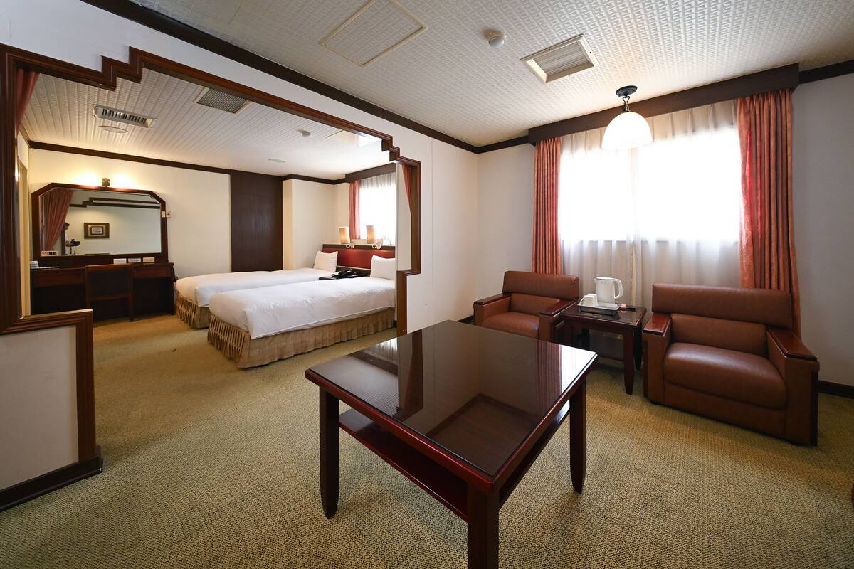 A hotel room with a bed, a desk, a chair, a coffee table, and a large window with curtains.