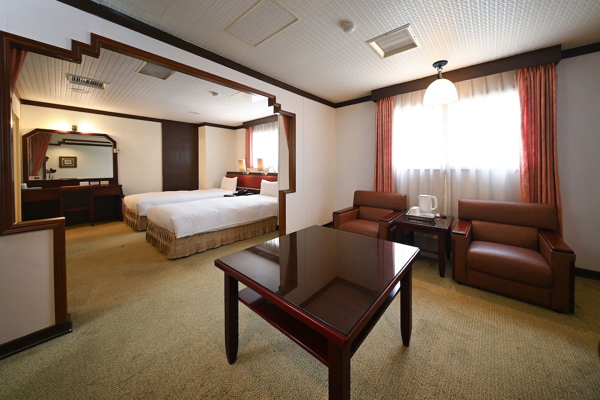 A hotel room with a bed, a desk, a chair, a coffee table, and a large window with curtains.