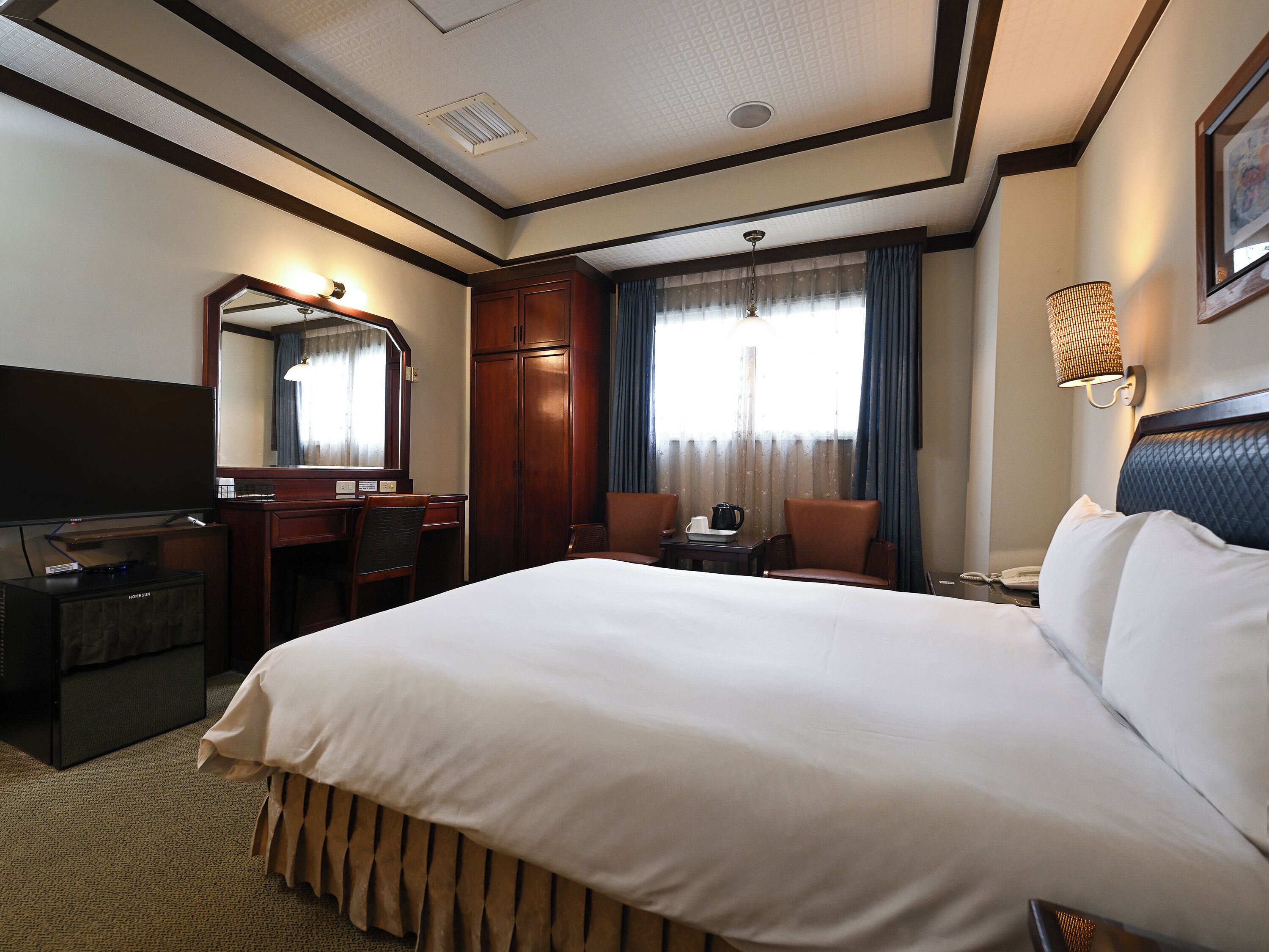 A hotel room with a bed, a desk with a chair, a television, a mirror, a window with curtains, and a wall-mounted lamp.