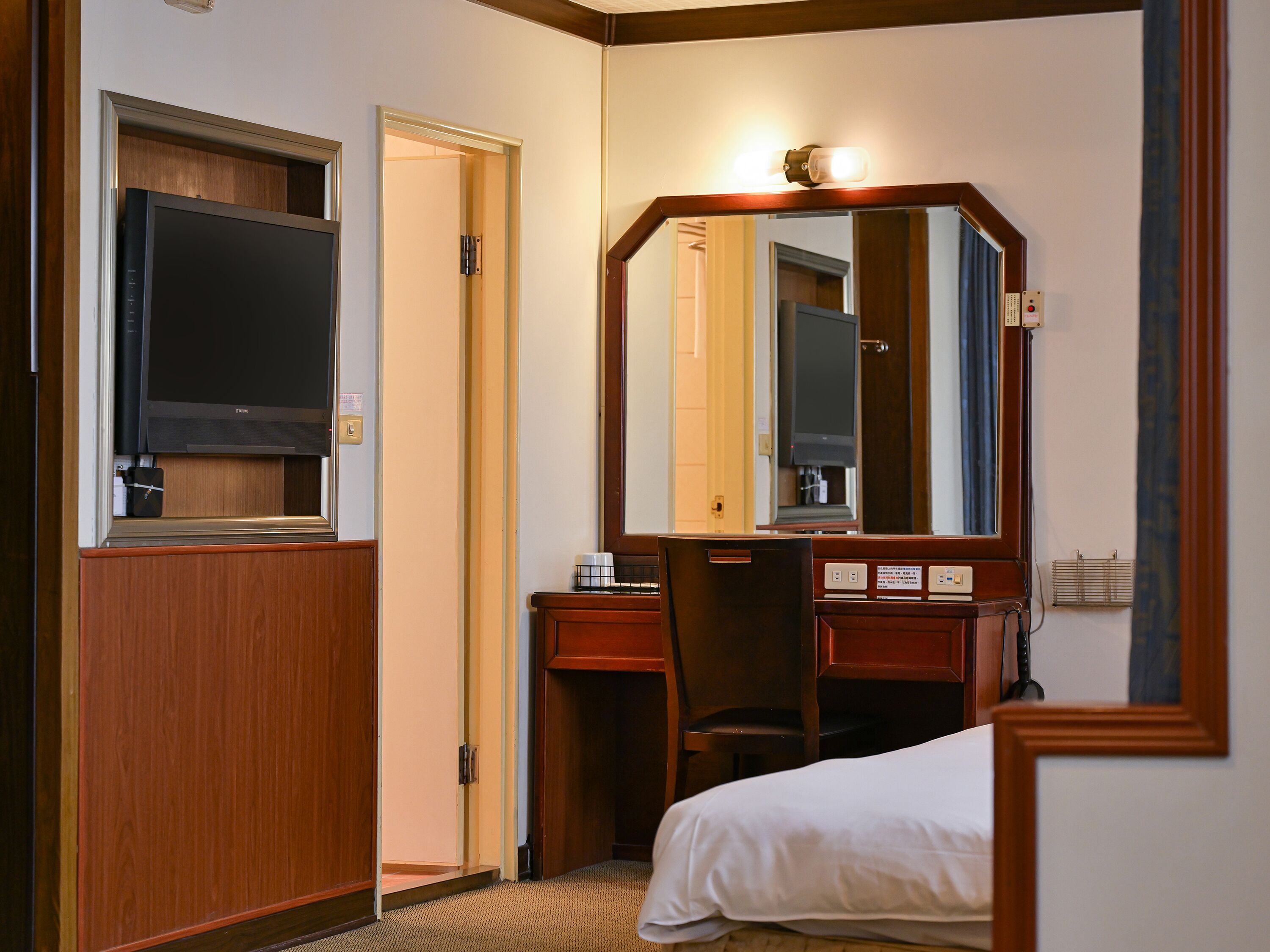 Habitación de hotel con una cama, un escritorio con silla, televisor, espejo y puerta.