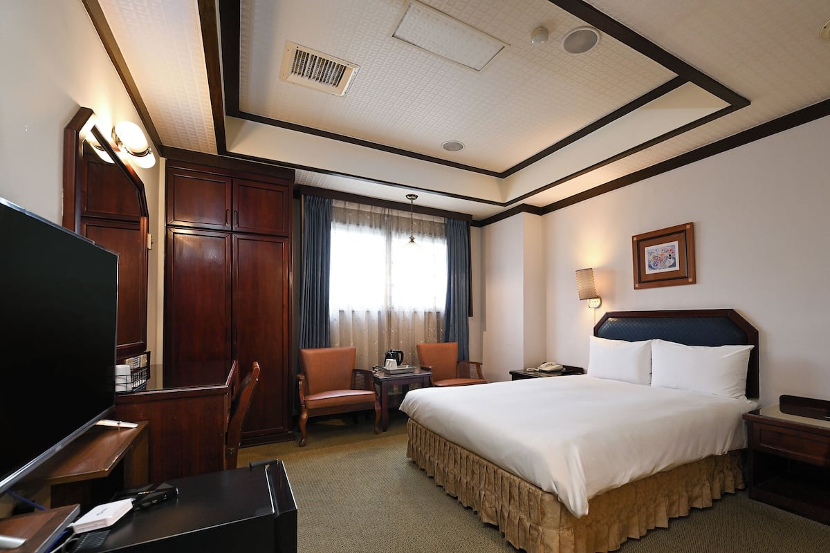 A hotel room with a bed, desk, chairs, a television, and a window with curtains.