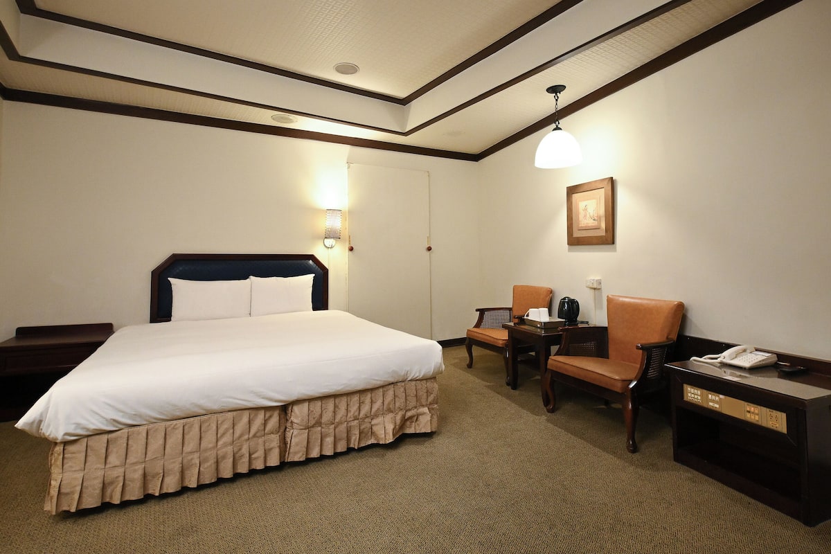 A hotel room with a bed, two chairs, a desk with a telephone, and a wall-mounted picture.