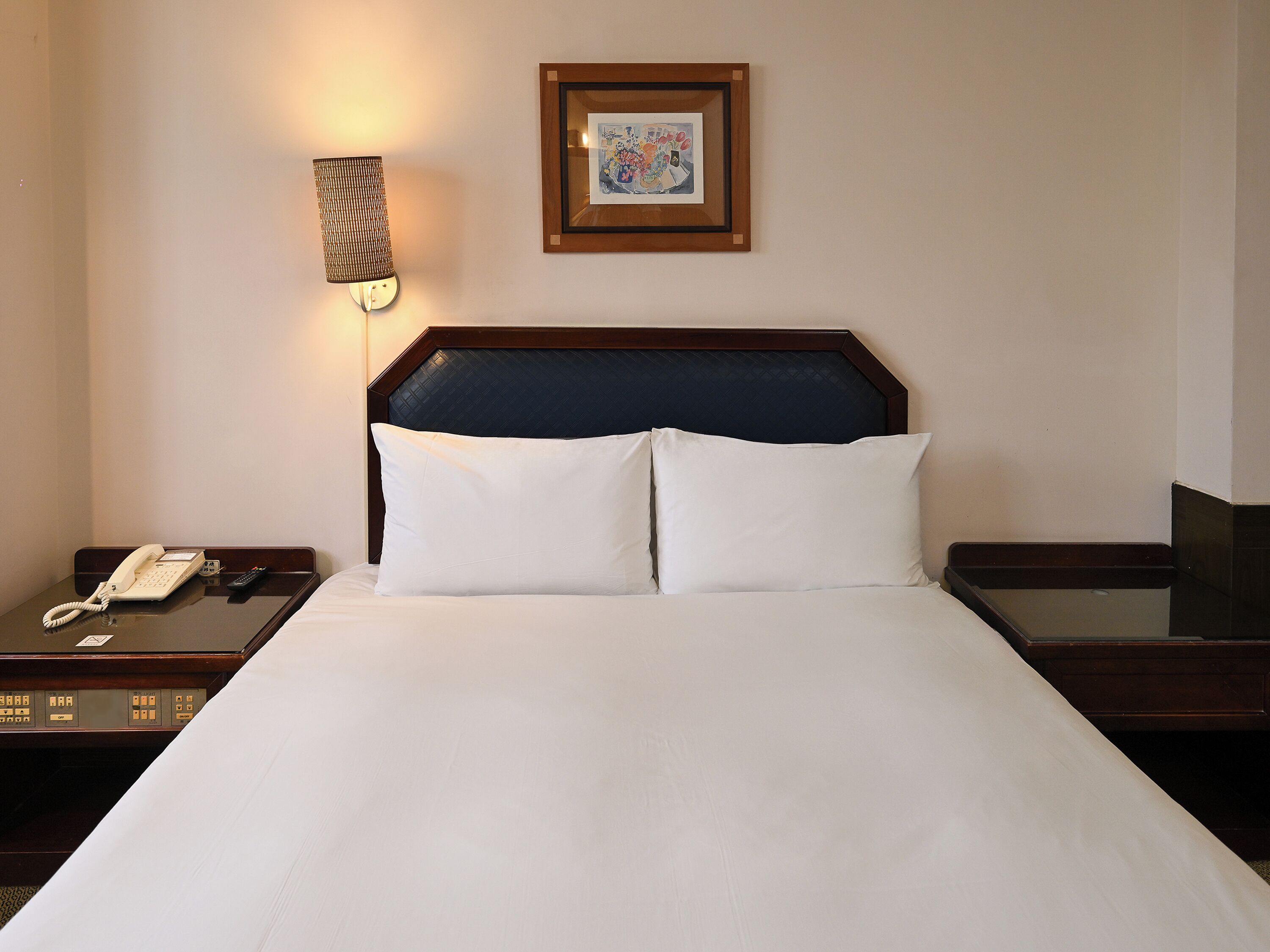 A hotel room with a bed, a bedside telephone, a framed picture on the wall, and a nightstand.