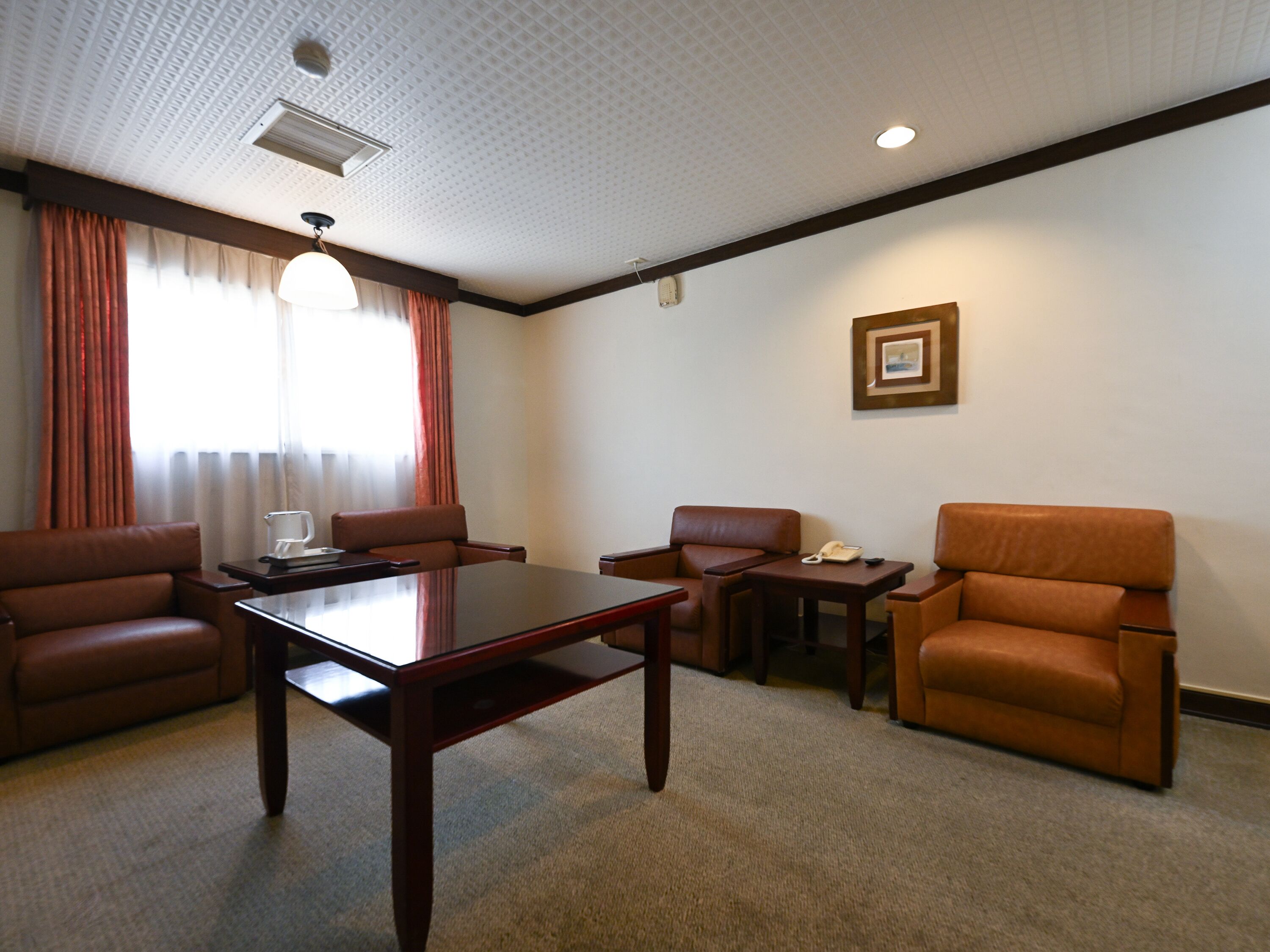 A hotel room with a wooden table, four leather chairs, a small side table, a framed picture on the wall, and a window with curtains.