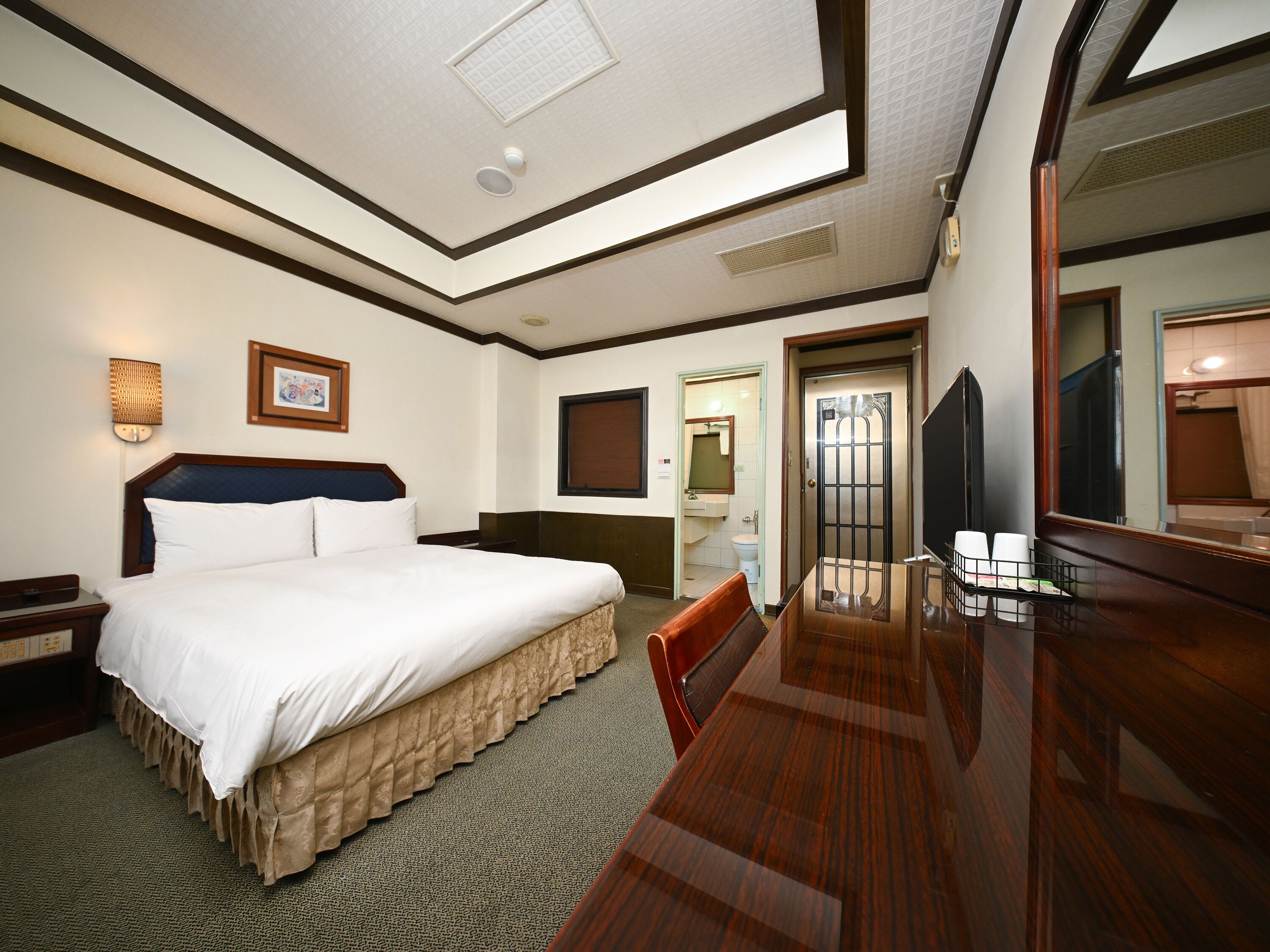 Quarto de hotel com uma cama, uma mesa, uma cadeira, um espelho e a porta do banheiro.