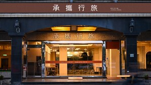 Entrada de un hotel con un letrero en caracteres chinos, fachada de vidrio y una banca en el exterior.