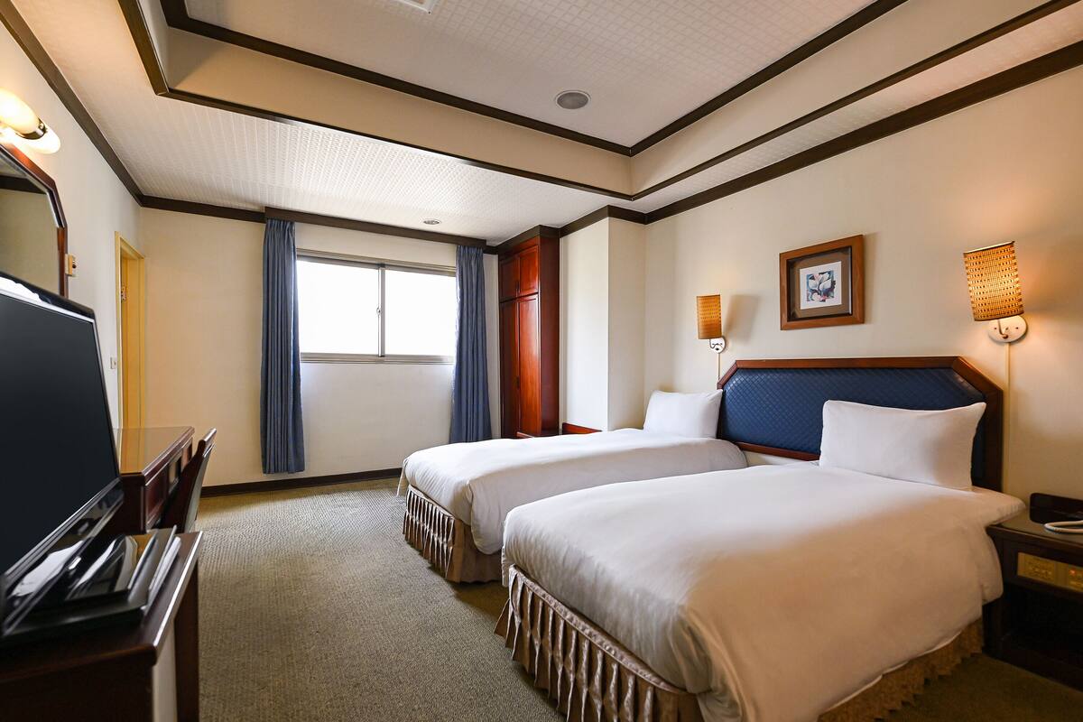 A hotel room with two beds, a desk, a television, and a window with curtains.