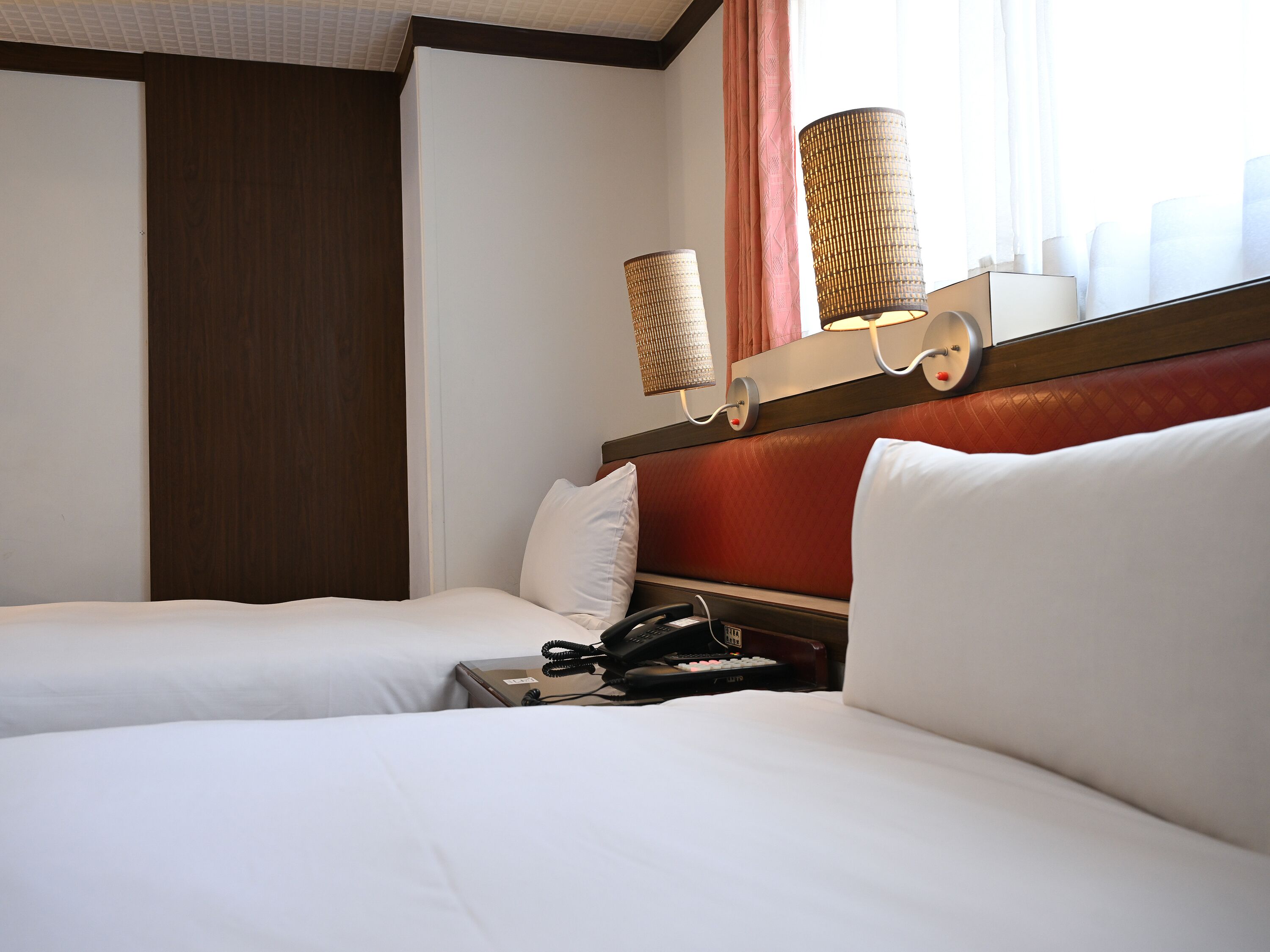 Quarto de hotel com duas camas, um telefone, uma luminária e uma janela com cortinas.
