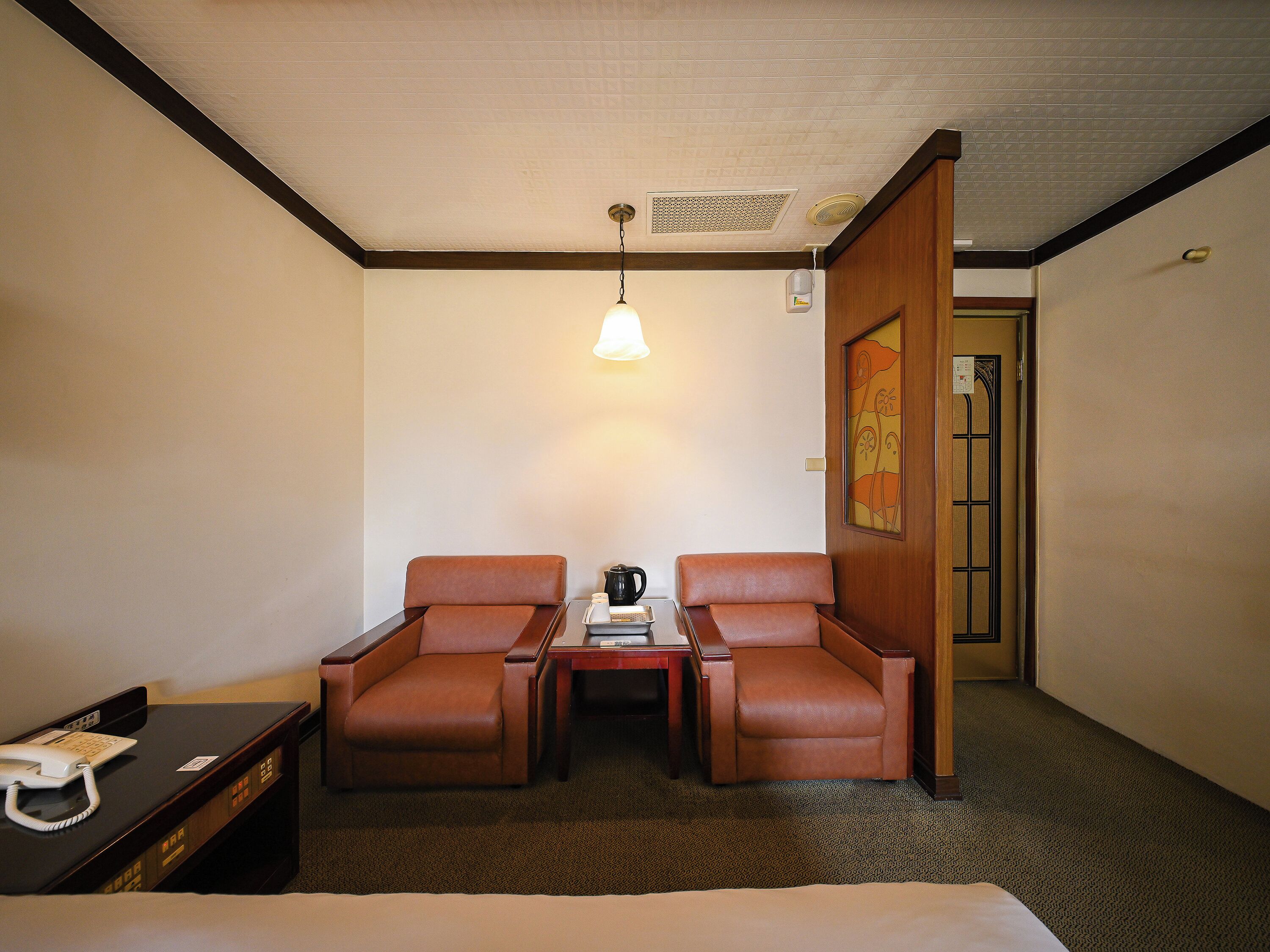 A hotel room with a bed, two brown leather chairs, a small table with a lamp, a telephone, and a door with a decorative panel.