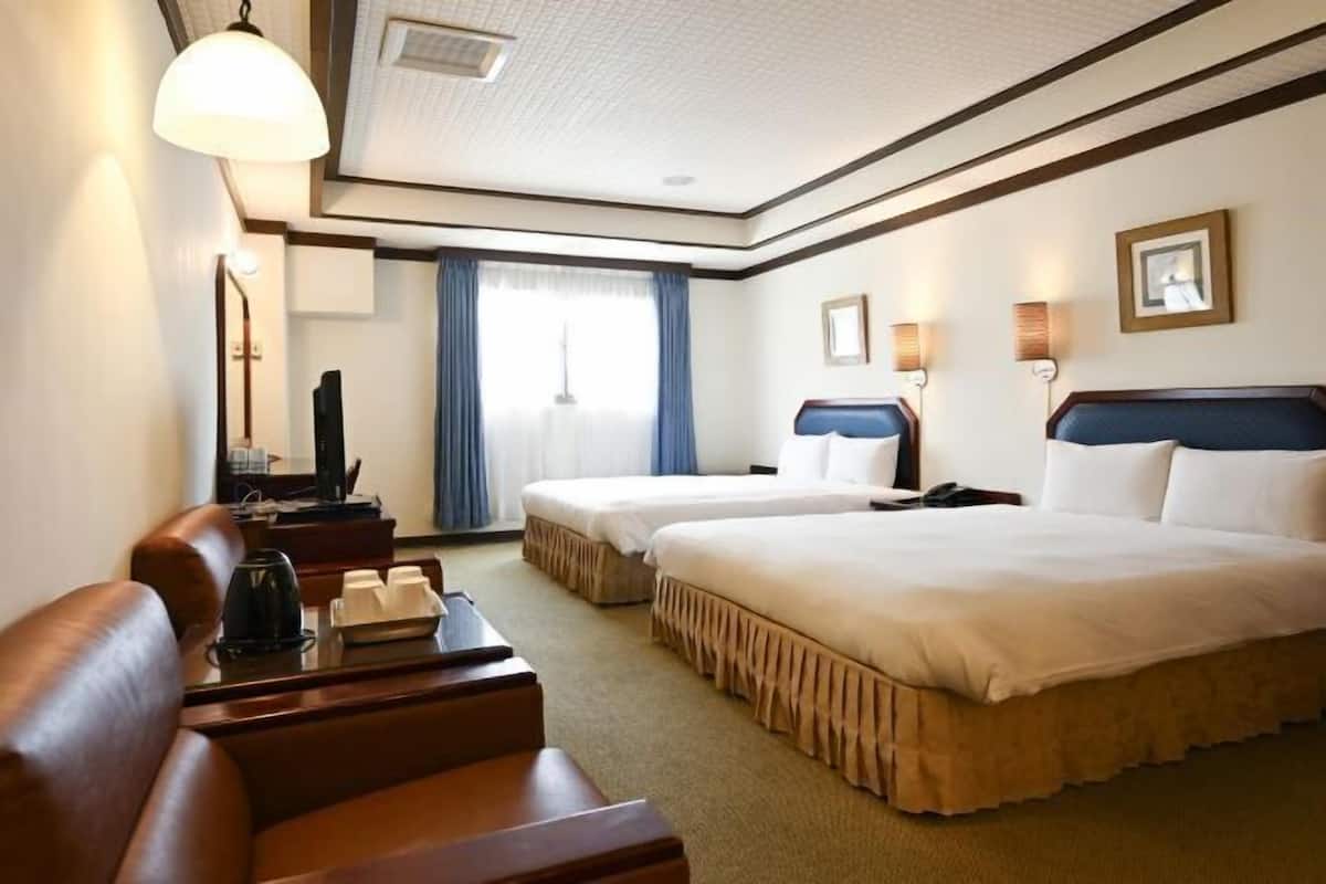 A hotel room with two beds, a desk, a chair, a TV, and a large window with curtains.