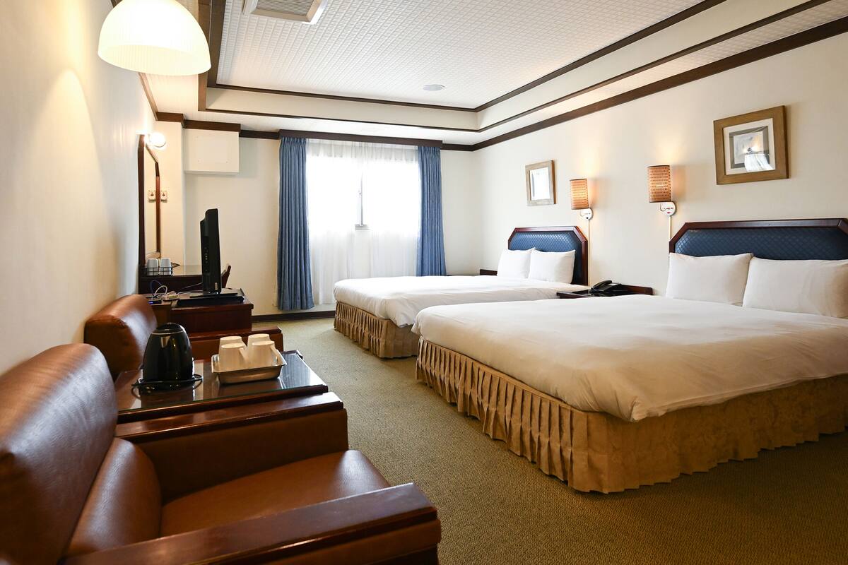 A hotel room with two beds, a sofa, a desk with a lamp, a TV, and a window with curtains.