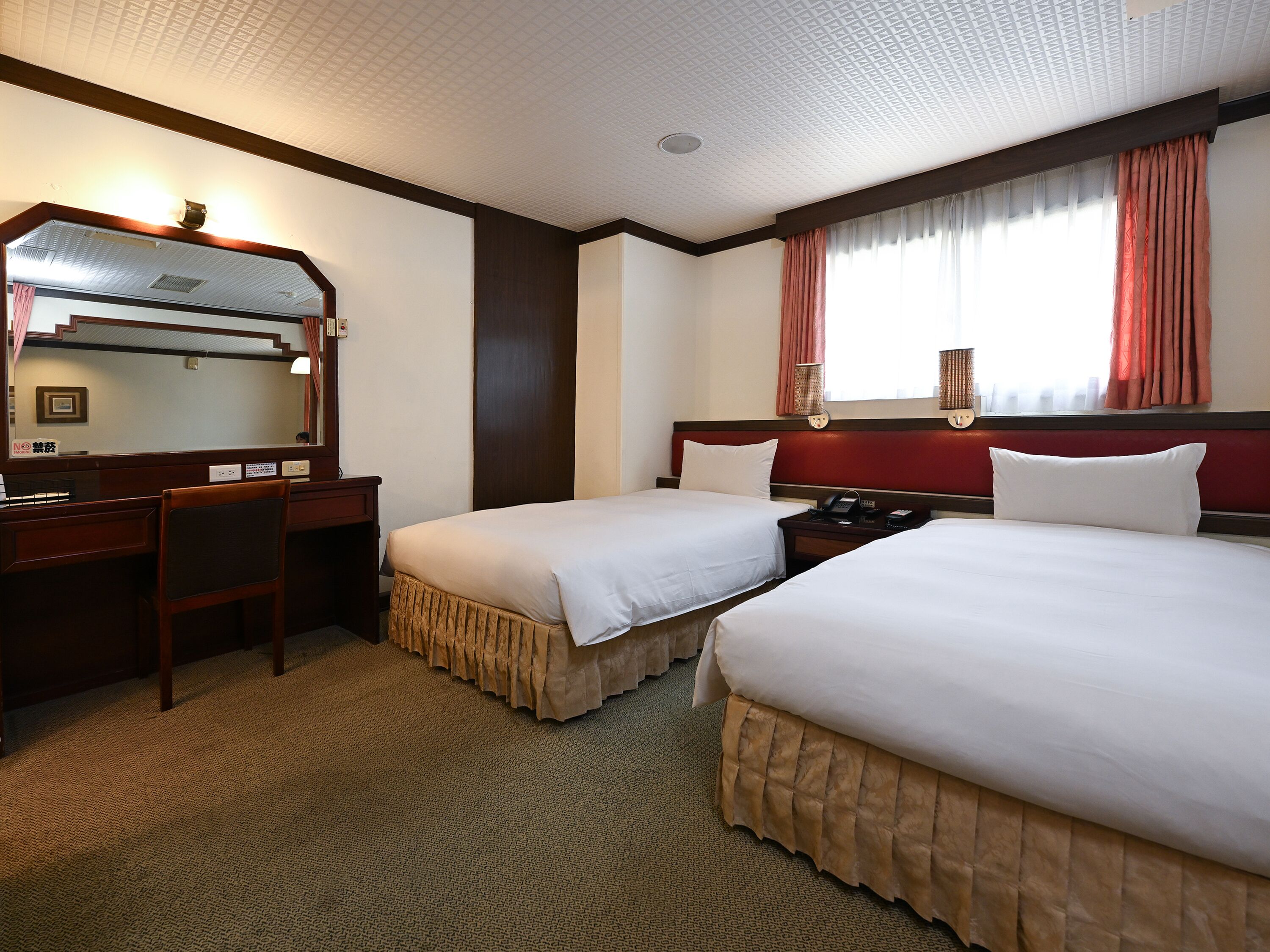 Habitación de hotel con dos camas, un escritorio, una silla, un espejo, un teléfono y una ventana con cortinas.