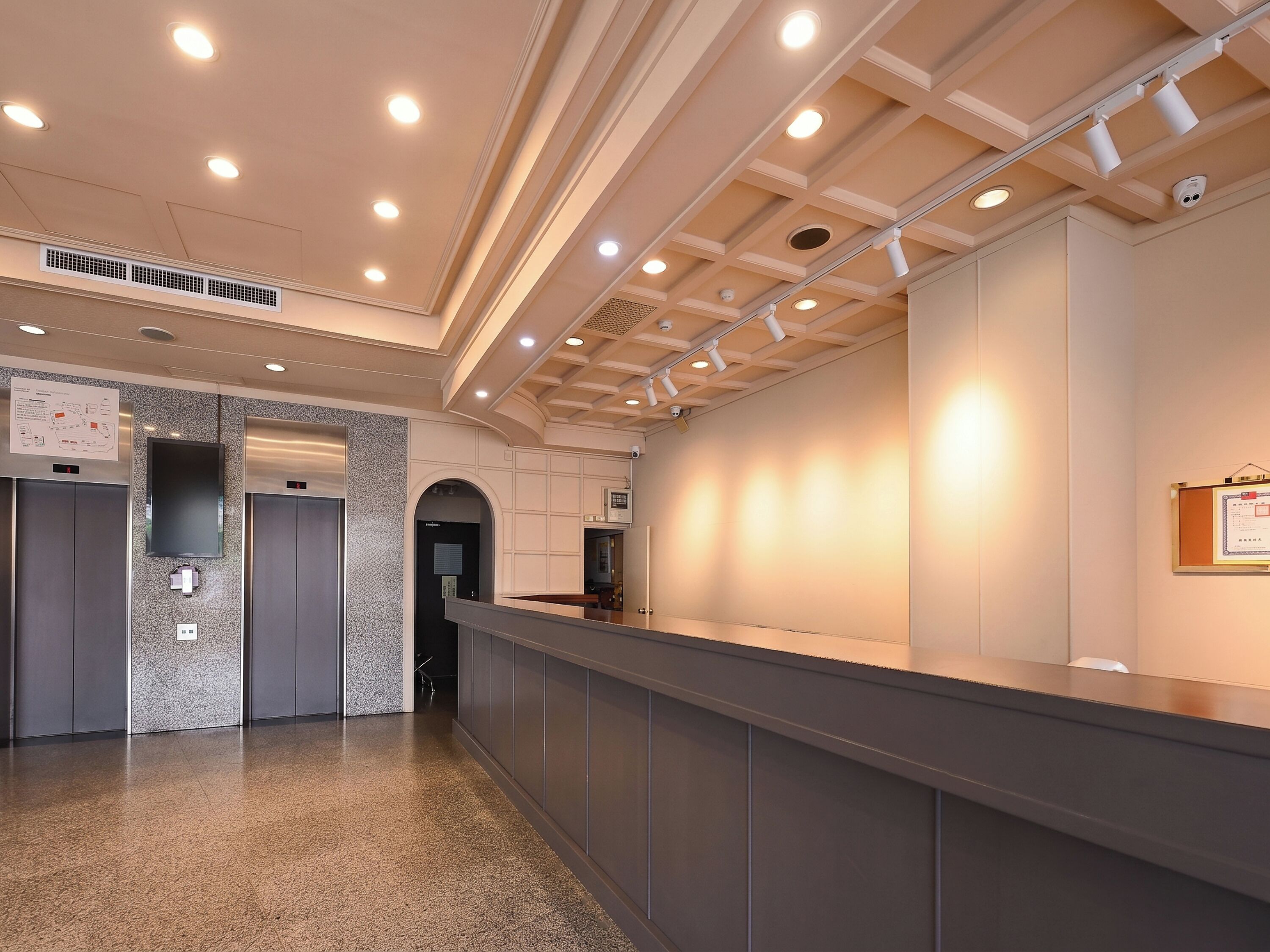 A modern lobby with a reception desk, elevators, a wall-mounted TV, and a ceiling with recessed lighting.