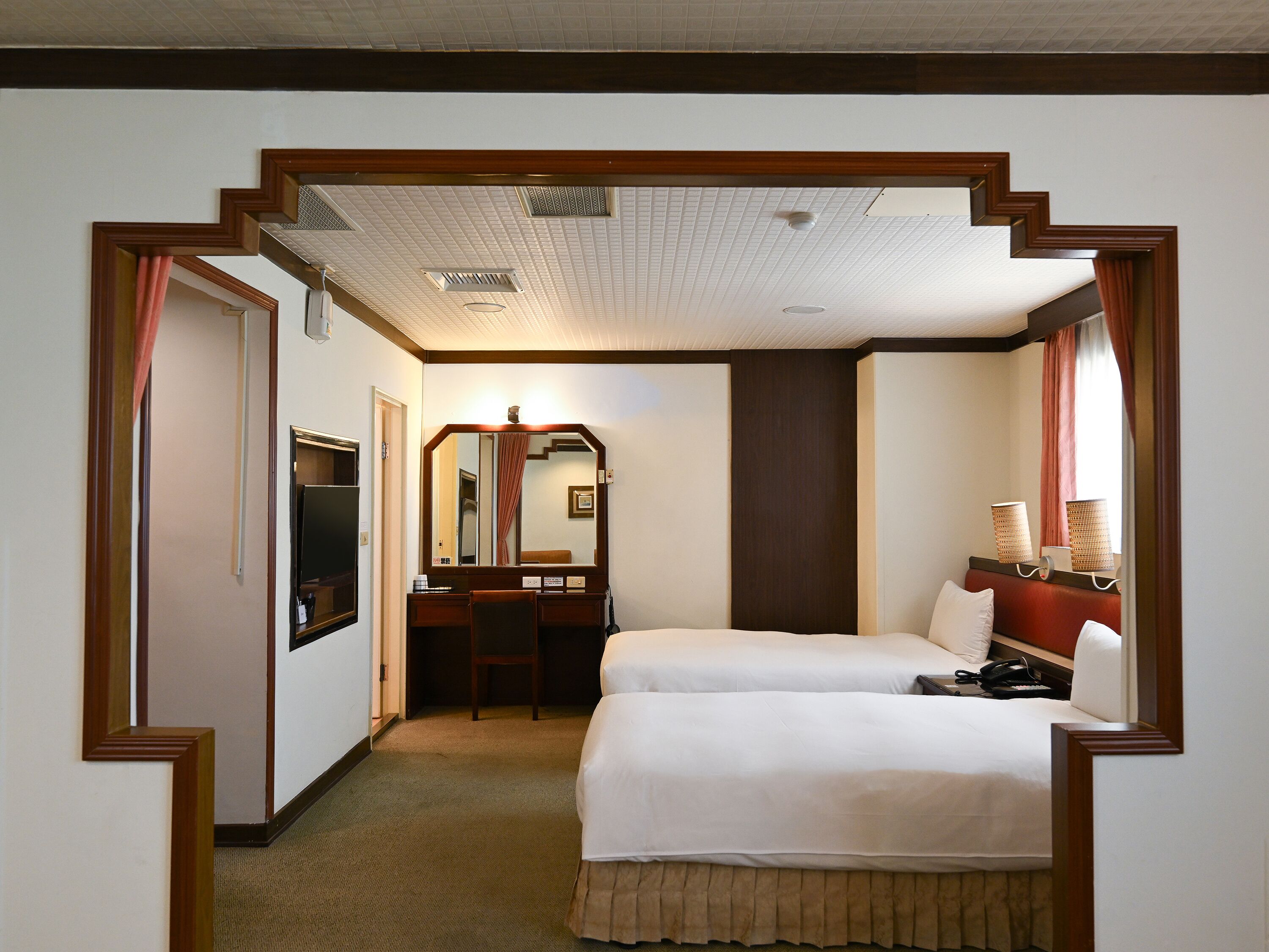 A hotel room with two beds, a desk, a mirror, a television, and a window with curtains.