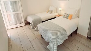 A room with two single beds, a nightstand with a lamp, a window with curtains, and a painting on the wall.