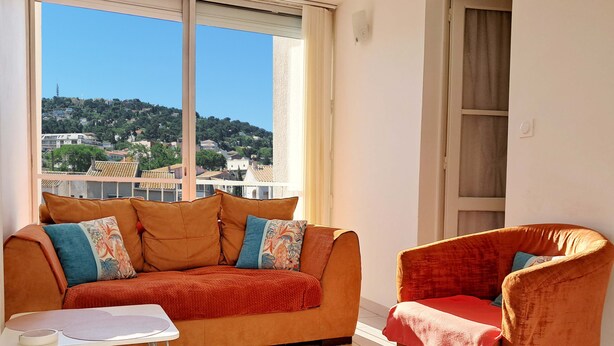 A room with a sofa and armchair, a small table, and a view of a hillside with buildings.