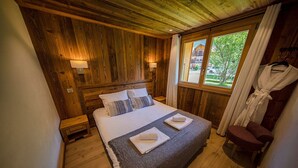 A wooden-paneled bedroom with a bed, bedside lamps, a window with curtains, and a robe hanging on the wall. - NEW Garden 7 person Apartment with Hot Tub, next to Super Morzine (Morzine)