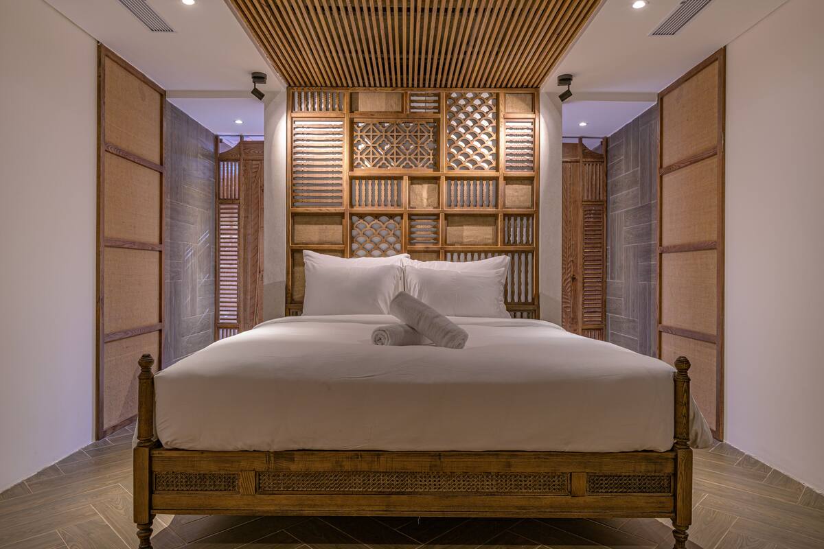 A large bed with a wooden frame and white bedding, set against a backdrop of a traditional wooden partition with intricate designs.