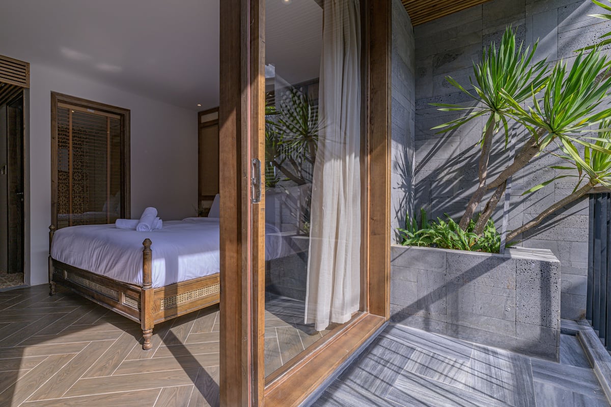 A bedroom with a wooden bed, a glass door leading to a balcony, and a view of a stone wall with plants.