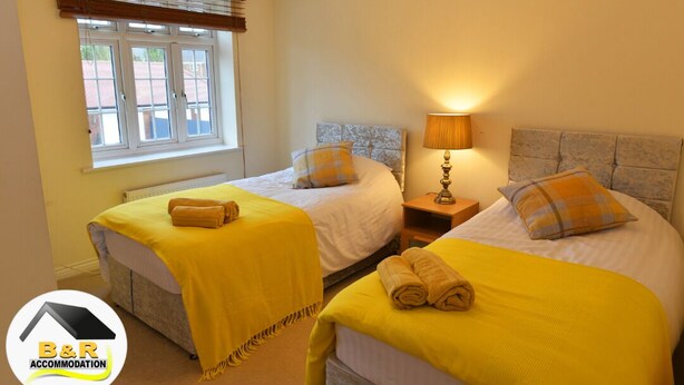 A hotel room with two beds, each with a yellow blanket and a pillow. There is a window with blinds, a bedside lamp, and a logo for B&R Accommodation. - ⭐B and R Serviced Accommodation and Apartments Amesbury⭐ Archer house  (Amesbury)
