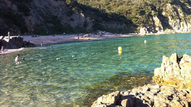 Una playa costera con aguas cristalinas, formaciones rocosas y un paisaje montañoso al fondo.