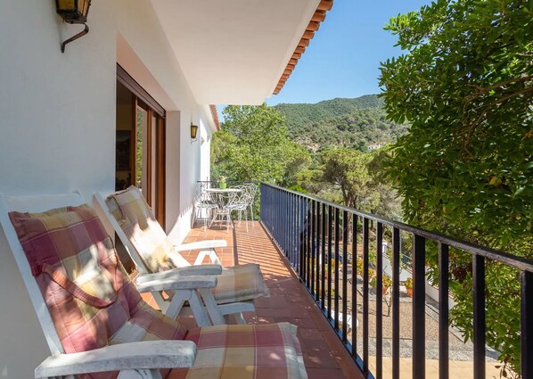A balcony with white lounge chairs and a table, overlooking a mountainous landscape.