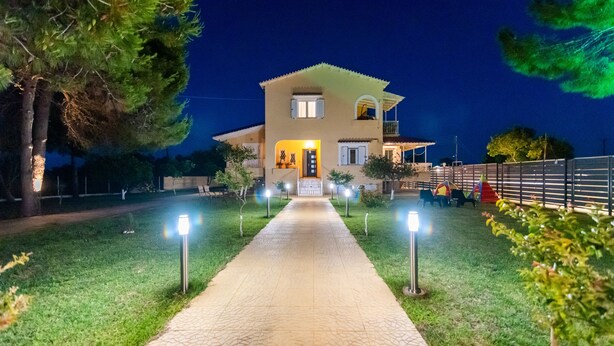 A two-story house with a paved pathway leading to the entrance, surrounded by greenery and illuminated by pathway lights. - Pine Trees Apartment (Preveza)