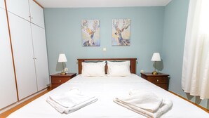 A neatly made bed with white linens and towels, two bedside tables with lamps, a wardrobe, and two wall-mounted paintings of deer. - Pine Trees Apartment (Preveza)