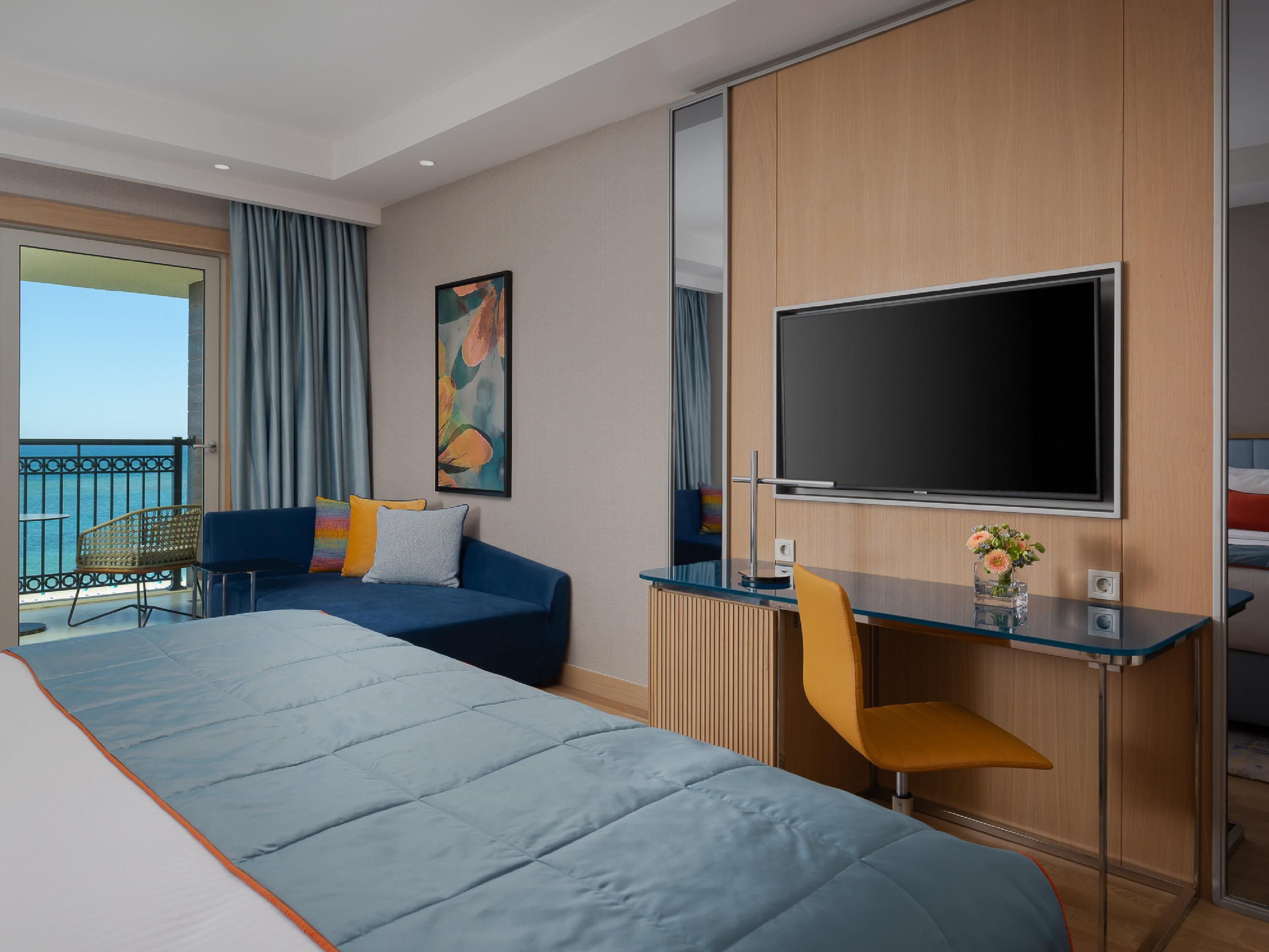 A modern hotel room with a large bed, a desk with a chair, a TV, and a balcony with a view of the sea.